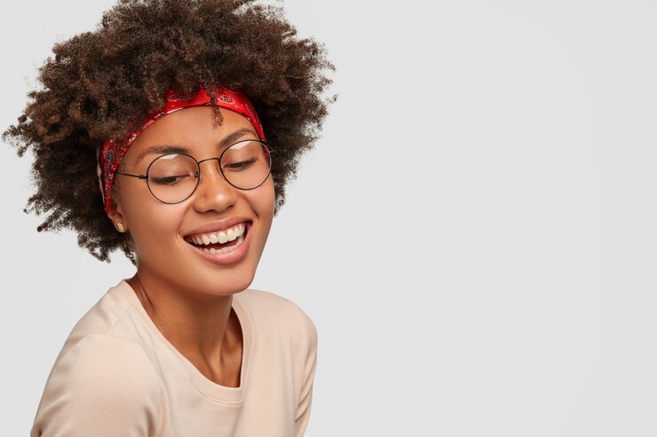 Photo gratuit charismatique heureuse jeune femme bouclée noire regarde vers le bas avec un large sourire, porte des lunettes transparentes, un bandeau rouge, rit de quelque chose de drôle, pose contre un mur blanc, espace copie vierge sur le côté droit.