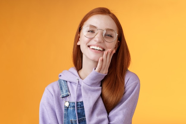 Charismatique heureuse rousse tendre souriante fille européenne des années 20 lunettes à capuche salopette en jean s'amusant à profiter des vacances d'été soirée fraîche en riant joyeusement toucher la peau claire et pure, fond orange.