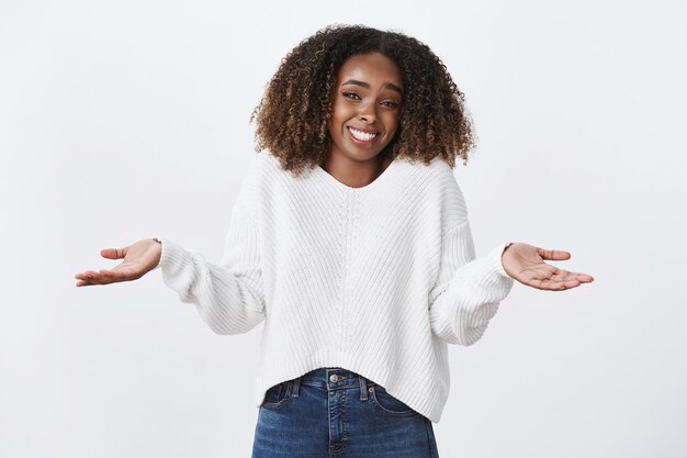 Charmant froid insouciant souriant femme afro-américaine coiffure frisée haussant les mains écartées sur le côté ne semble pas dérangé ne donnant pas d'intérêt, debout sans aucune idée, mur blanc inconscient