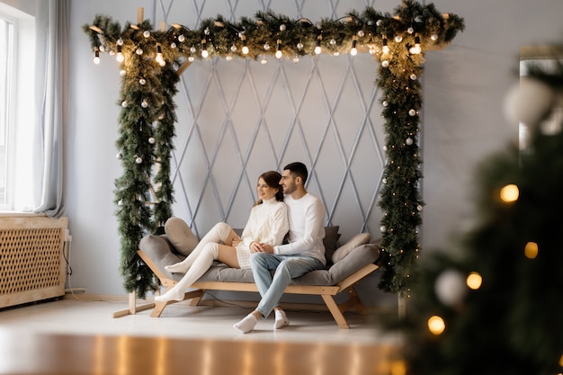 Charmant jeune couple en vêtements de maison blancs confortables pose dans une chambre avec arbre de Noël