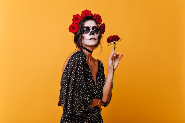 Charmant modèle féminin en robe élégante appréciant le parfum des roses. Portrait de jeune fille avec du maquillage d'Halloween posant dans la salle orange.