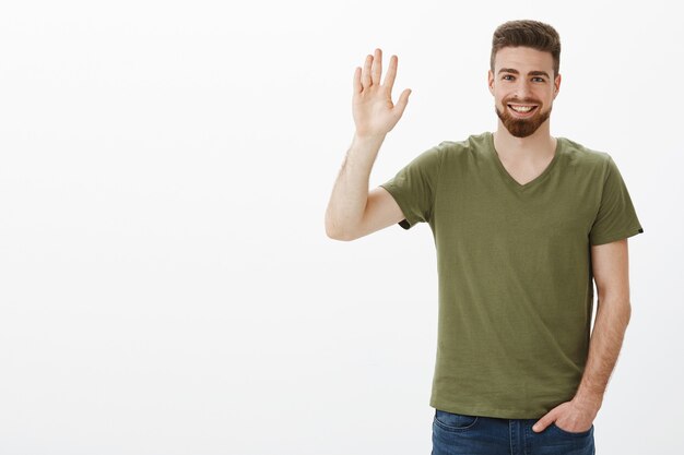 Charmant petit ami barbu d'apparence amicale se sentant timide en agitant la main levée en bonjour ou salut geste, tenant la main dans la poche avec désinvolture en rencontrant de nouvelles personnes, saluant sur un mur blanc