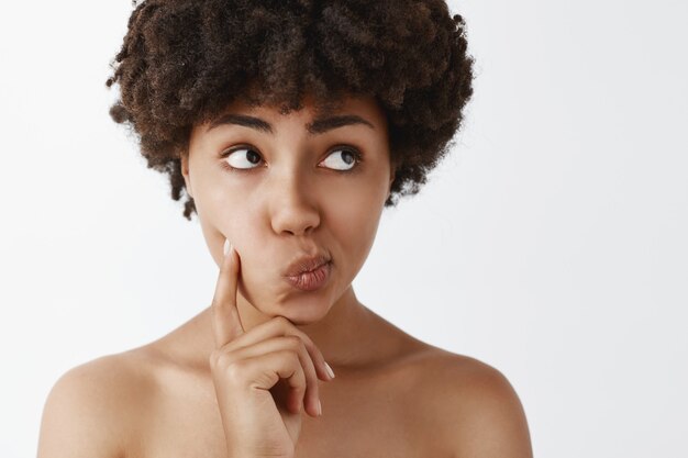 Charmante afro-américaine nue tendre et féminine aux cheveux bouclés, toucher la joue, tourner les lèvres à droite et lever les yeux tout en pensant ou en décidant quelque chose