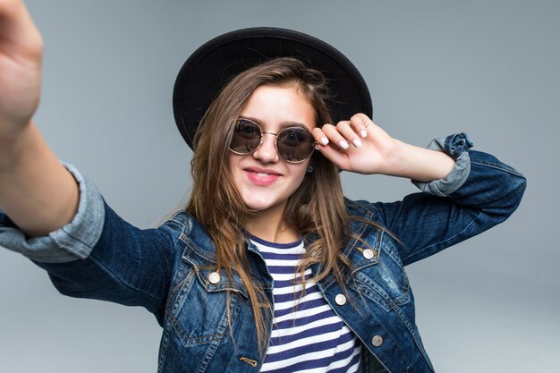 Charmante belle femme au chapeau noir et lunettes de soleil prendre selfie des mains sur fond gris