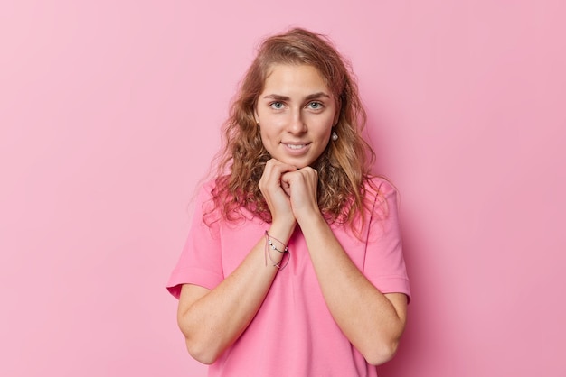 Charmante belle jeune femme aux yeux bleus avec de longs cheveux ondulés garde les mains sous le menton regarde directement la caméra vêtue d'un t-shirt de base décontracté isolé sur fond rose pose pour faire de la photo