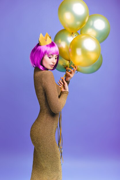 Charmante belle jeune femme en robe à la mode attrayante avec des ballons dorés volants. Coupe de cheveux violet rose, couronne, émotions joyeuses, yeux fermés, célébration.