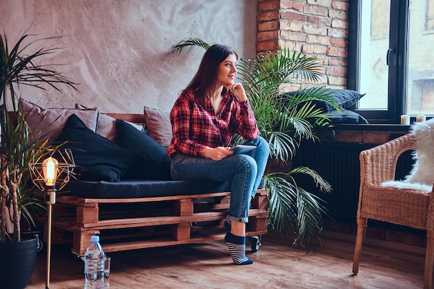 Charmante brune dans une chemise en flanelle et un jean assis sur un canapé