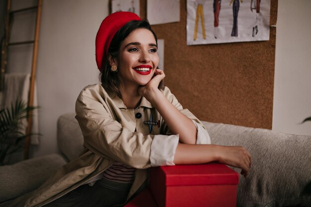 Charmante dame en béret rouge élégant souriant et assis sur le canapé Magnifique jeune femme aux cheveux noirs en manteau beige posant