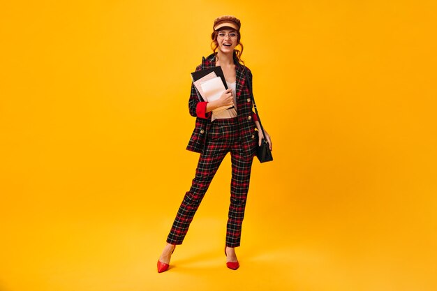 Charmante dame en costume à carreaux et casquette pose avec des cahiers sur un mur orange