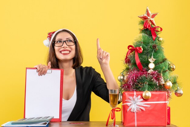 Charmante dame en costume avec chapeau de père Noël et lunettes montrant le document pointant vers le haut dans le bureau sur jaune isolé