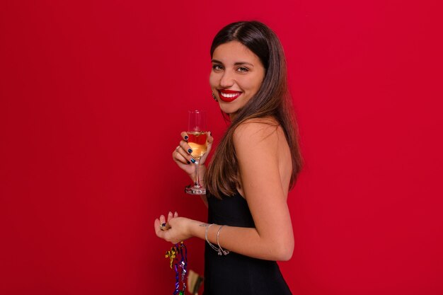 Charmante dame avec grand sourire posant sur un mur rouge avec une coupe de champagne se préparant pour la fête de Noël