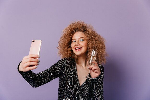 La charmante dame en haut noir brillant rit, fermant les yeux, tenant un verre de champagne et faisant un selfie sur un espace violet.