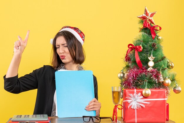 Charmante dame réfléchie en costume avec chapeau de père Noël et décorations de nouvel an tenant un document dans le bureau sur isolé jaune