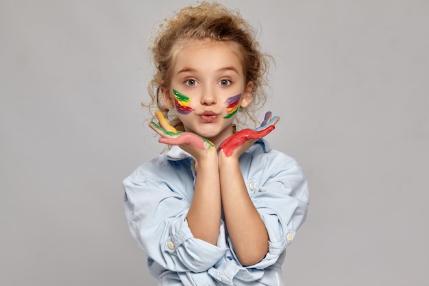 Photo gratuite charmante écolière ayant une brosse dans ses cheveux blonds bouclés chics, vêtue d'une chemise bleue et d'un t-shirt blanc. elle a peint ses joues et a donné un baiser à la caméra sur un fond gris.