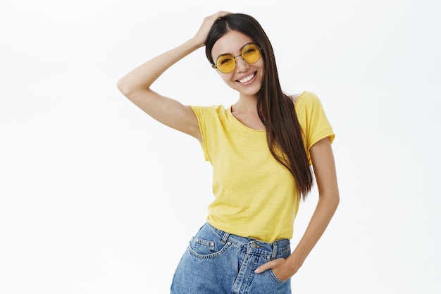 Charmante et élégante photographe indépendante insouciante dans des lunettes de soleil à la mode et un t-shirt jaune touchant les cheveux