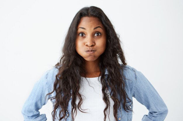 Charmante femme afro-américaine moqueuse portant des cheveux ondulés lâches à la recherche avec les yeux sur écoute avec la bouche pleine. Modèle à peau foncée posant sur un mur blanc isolé.