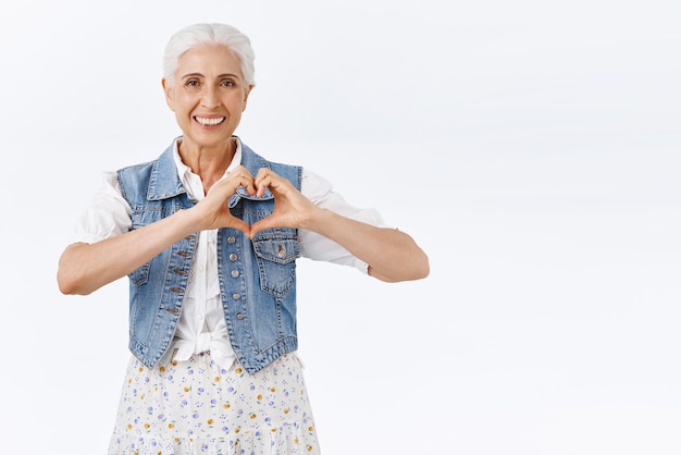 Charmante femme âgée caucasienne vivante et insouciante vieille dame montrant le signe du coeur exprimer l'amour ou les soins souriant heureux chérir la famille et les relations chaleureuses comme passer du temps avec les petits-enfants