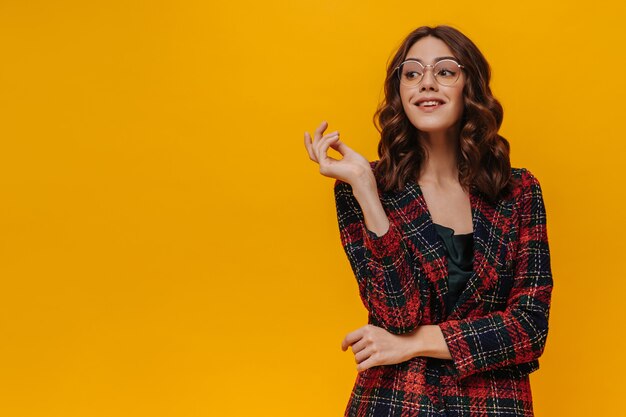 Charmante femme aux cheveux bouclés dans des lunettes posant sur un mur isolé