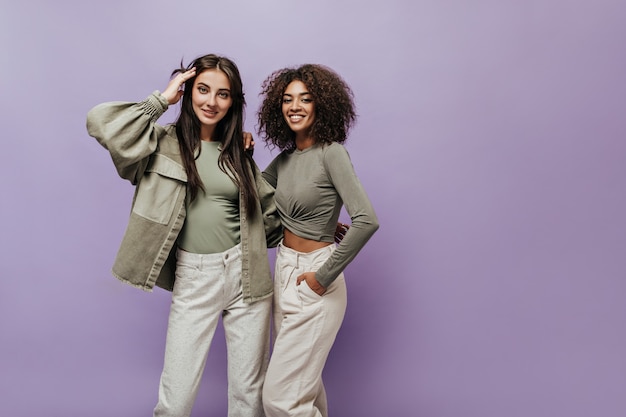 Charmante femme aux cheveux bouclés en haut olive et pantalon blanc souriant et étreignant avec une fille élégante en veste sur un mur lilas