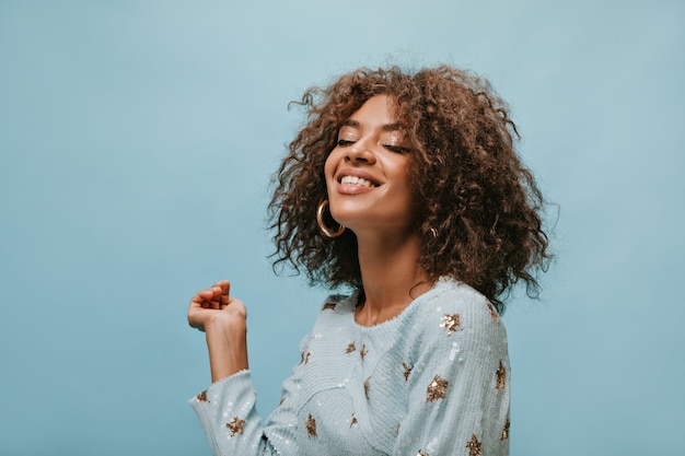 Charmante femme aux cheveux courts brune dans des boucles d'oreilles en or élégantes et des vêtements bleus imprimés souriant les yeux fermés sur un mur isolé