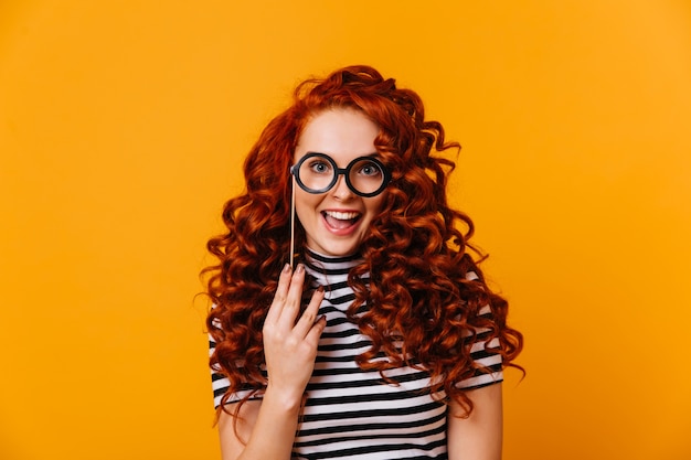 Charmante femme aux yeux bleus et aux cheveux rouges tient le modèle de lunettes et sourit sur l'espace orange.