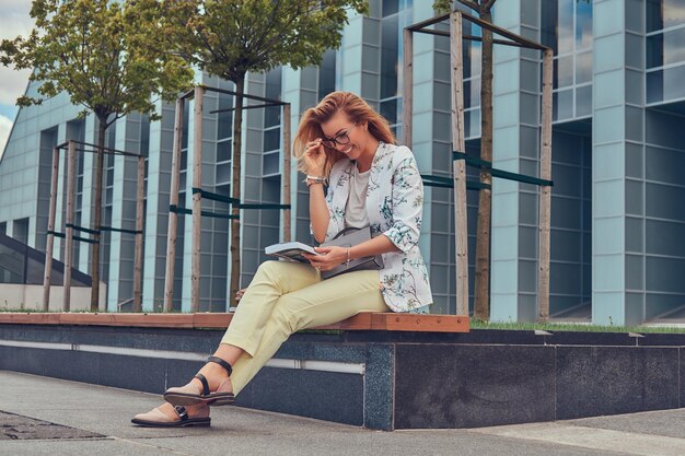 Charmante femme blonde en vêtements modernes, étudiant avec un livre, assise sur un banc dans le parc contre un gratte-ciel