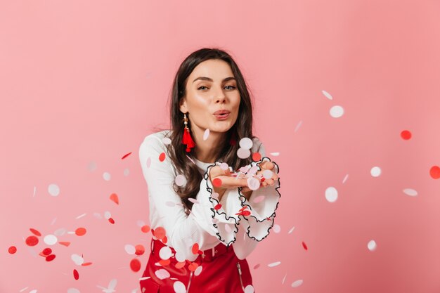 Une charmante femme de bonne humeur souffle des confettis rouge-blanc de ses paumes. Fille en boucles d'oreilles massives posant sur fond isolé.