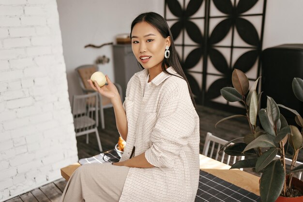 Charmante femme brune bronzée en cardigan beige et pantalon se penche sur la caméra sourit et tient la pomme Jolie dame asiatique est assise sur un bureau en bois dans la cuisine