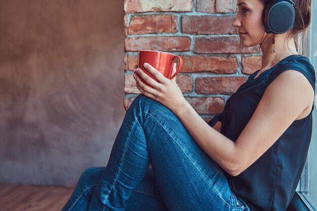 Charmante femme brune en chemisier noir écoutant de la musique dans des écouteurs et tenant une tasse de café tout en étant assise sur un rebord de fenêtre dans une pièce avec un intérieur loft.