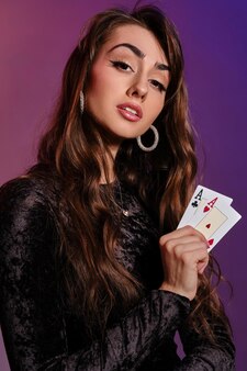 Charmante femme brune avec maquillage, en robe de velours noir et bijoux montre deux cartes à jouer et vous regarde, posant sur un fond coloré. jeux d'argent, poker, casino. fermer.