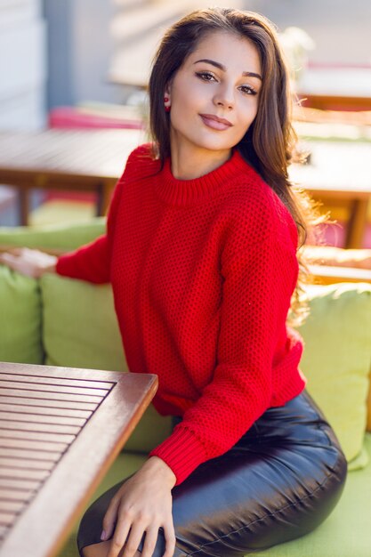 Charmante femme brune en pull tricoté automne rouge et jupe en cuir reposant sur un canapé dans un restaurant en espace ouvert.