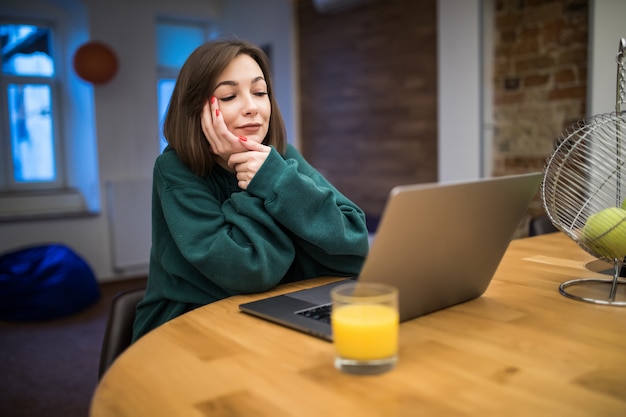 Charmante femme brune travaille sur son ordinateur portable sur la table de la cuisine, boire du jus d'orange