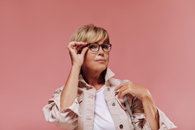 Charmante femme avec une coiffure moderne courte et des lunettes dans une veste légère élégante regardant dans la caméra sur fond rose isolé