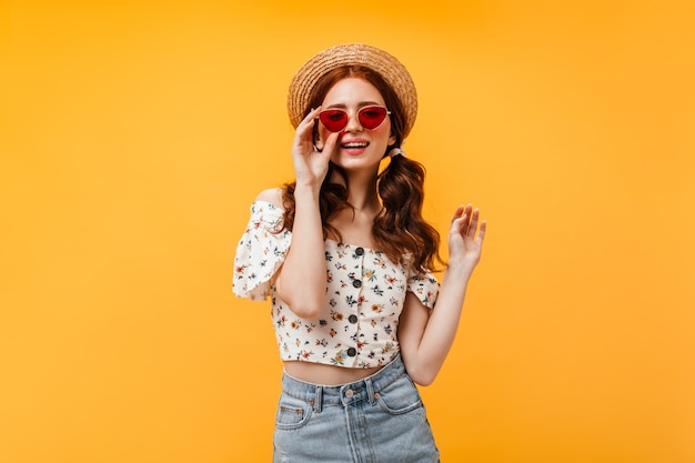 Charmante Femme Frisée En Chemisier Court élégant Et Jupe En Jean Porte Des Lunettes De Soleil Rouges. Femme En Plaisancier Se Penche Sur La Caméra Sur Fond Orange.