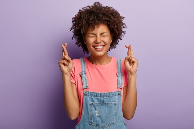 Charmante femme heureuse attend le rêve devenu réalité, croise les doigts pour la bonne chance, a le sourire à pleines dents, les yeux fermés, vise le succès