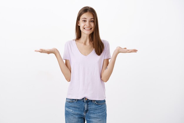 Charmante femme heureuse à la recherche amicale avec de courts cheveux bruns sprading paumes gauche et droite comme si l'objet sur l'espace de copie contre le mur gris