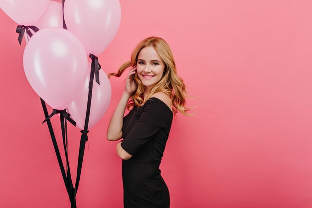 Charmante femme mince jouant avec ses cheveux blonds debout près des ballons. Photo intérieure d'une charmante fille de joyeux anniversaire se détendre pendant la fête.