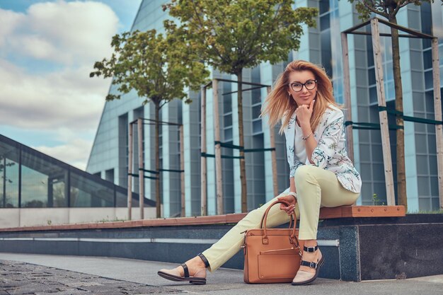 Charmante femme à la mode dans des vêtements élégants et des lunettes avec un sac à main, assise sur un banc contre un gratte-ciel.