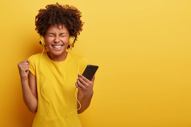 Charmante femme à la peau sombre satisfaite tient le smartphone, serre le poing, ferme les yeux du plaisir