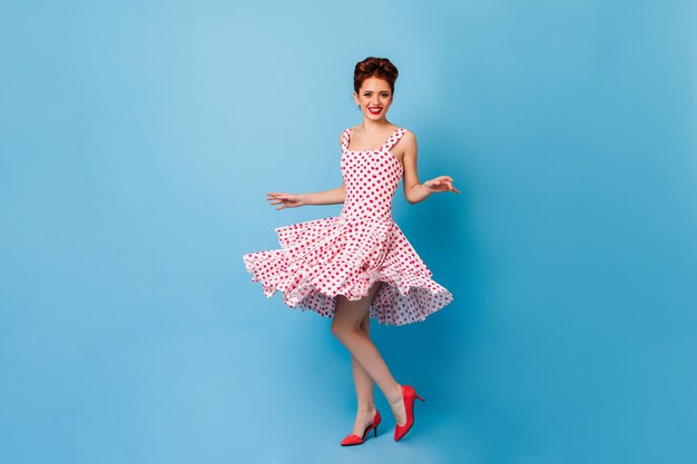 Charmante femme pin-up regardant la caméra avec un sourire sincère. Photo de Studio d'un modèle féminin en robe à pois dansant sur l'espace bleu.