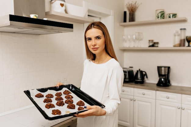 Une charmante femme positive en vêtements blancs tient des biscuits du four et se pose devant la caméra avec le sourire à la cuisine de la maison