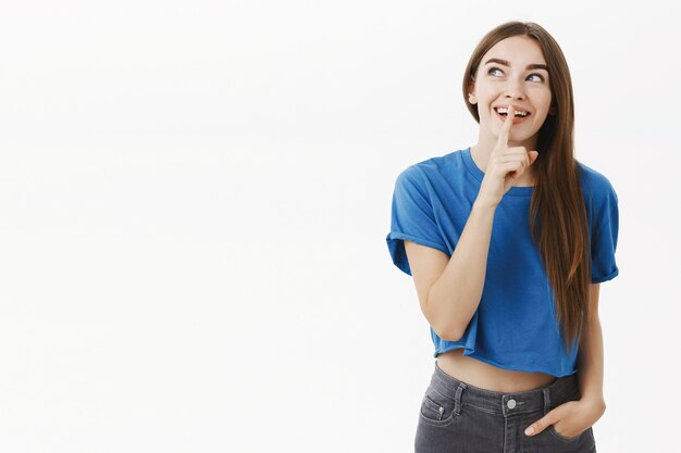 Charmante femme rêveuse et amusée en t-shirt bleu regardant excité dans le coin supérieur gauche, souriant largement montrant un geste chut avec l'index sur la bouche en gardant le secret ou en faisant la surprise