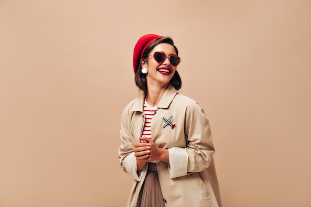 Charmante femme en robe beige et béret rouge rit sur fond isolé. Jolie jeune femme à lunettes de soleil et avec des lèvres brillantes posant à la caméra.