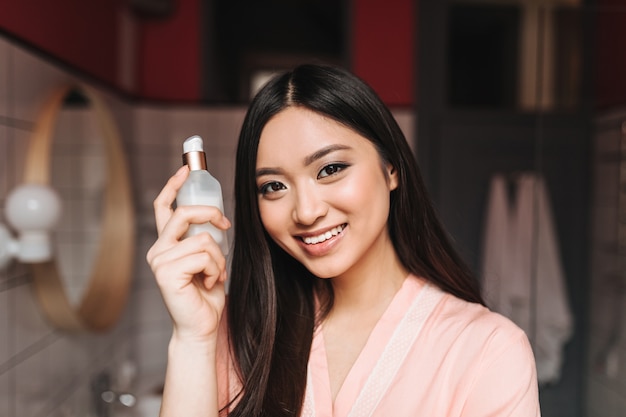 Charmante femme en robe de chambre en soie légère tient un pot de sérum pour le visage et souriant