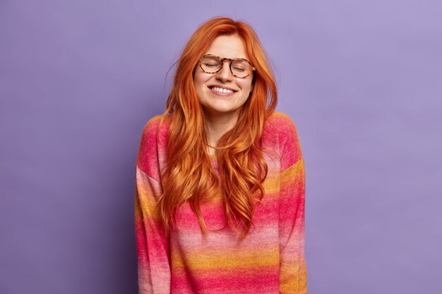 Charmante femme rousse heureuse se tient les yeux fermés et un large sourire rappelle un moment agréable de la vie porte un pull confortable.