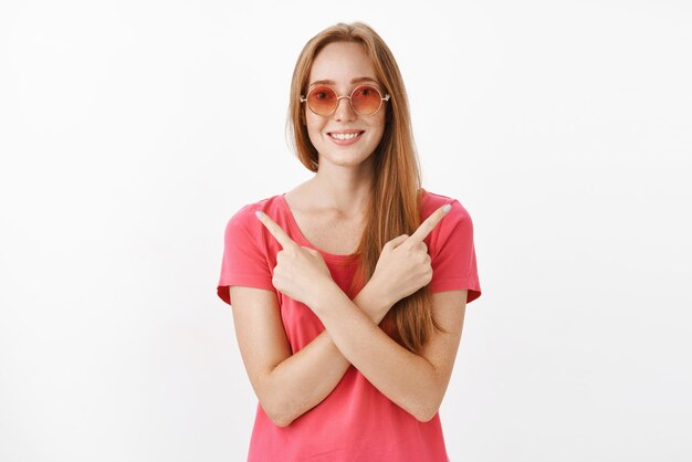 Charmante femme sans soucis avec des cheveux roux et des taches de rousseur dans des lunettes de soleil à la mode croisant les mains sur le corps pointant à gauche et à droite en étant sûr que les deux décisions sont bonnes sur un mur blanc