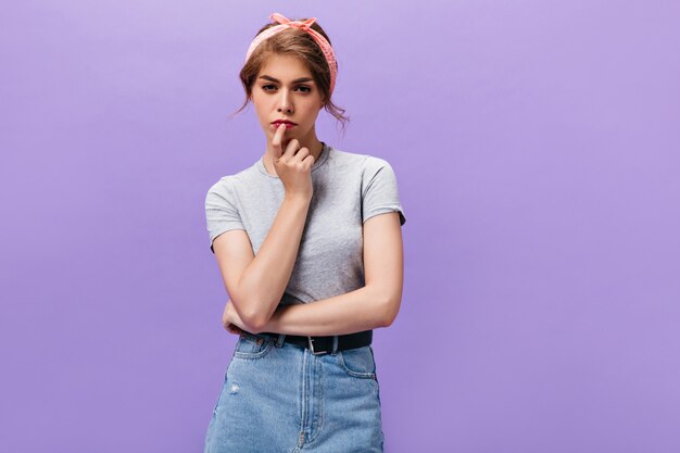 Charmante femme en T-shirt gris et jupe en jean pose pensivement sur fond isolé. Belle fille en tenue d'été à la recherche dans la caméra.