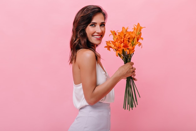 Charmante femme en tenue blanche sourit et tient des fleurs orange. Jolie dame adulte en longue robe d'été regarde la caméra et sourit.