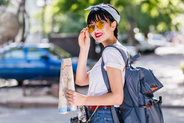 Charmante fille aux lèvres rouges tient de manière ludique des lunettes jaunes et souriant pendant un voyage autour de la ville avec sac à dos