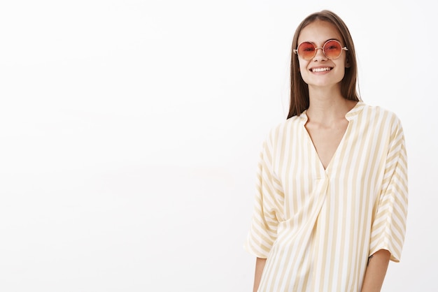 Charmante fille bénéficiant d'une journée ensoleillée lwarm marchant le long de la plage en chemisier à rayures jaune à la mode et lunettes de soleil souriant joyeux